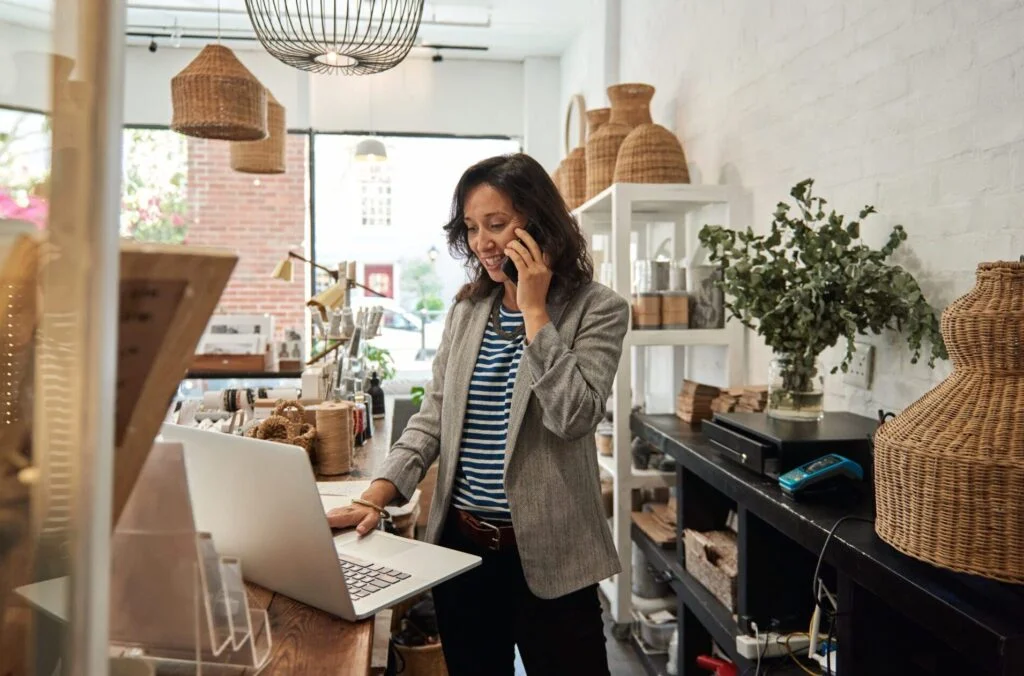Smiling Asian woman talking on a cellphone in her store Smiling young Asian woman standing behind a counter in her stylish boutique working on a laptop and talking on a cellphone