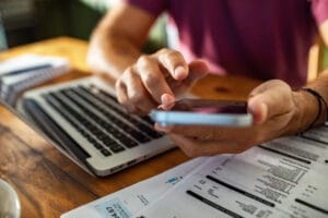 Man checking phone while reviewing bills and using laptop at kitchen table Man checking phone and using laptop at kitchen table to spot errors on utility bills