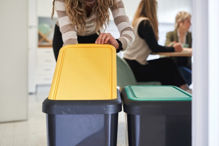 Woman recycling waste in office kitchen with color-coded bins representing waste management