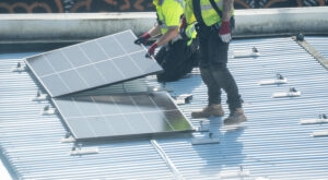 Two worker in yellow vest installing solar panel on metal rooftop surface Two technicians placing solar panel on metal roof in bright daylight