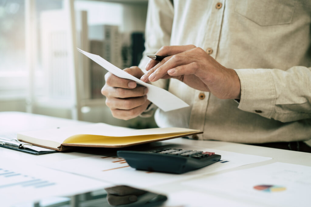 Deduction planning, debt asian accountant hand using calculator to calculating money balance from bill and income, cost budget expenses for pay money form personal individual on table at home. man with receipt and pen surrounded by calculator etc representing VAT check